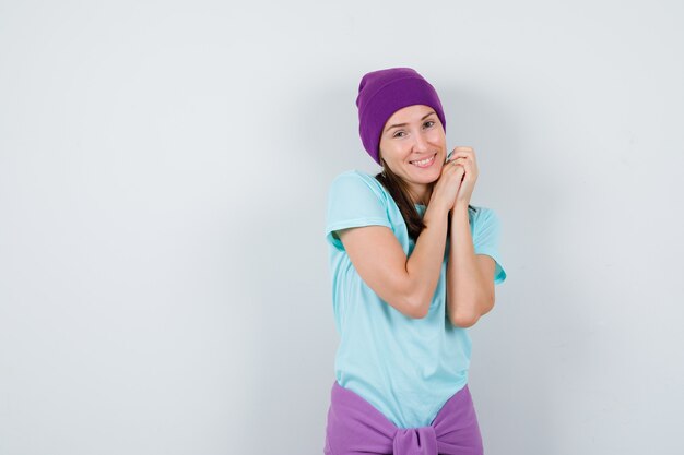 Young woman posing with clasped hands in t-shirt, beanie and looking joyful. front view.