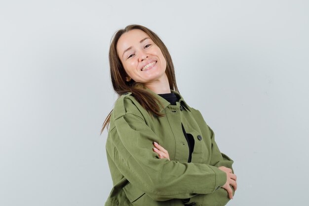 Young woman posing with crossed arms in green jacket and looking jolly , front view.