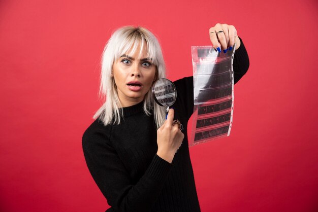 Young woman posing with a loupe and a photo tape on a red background