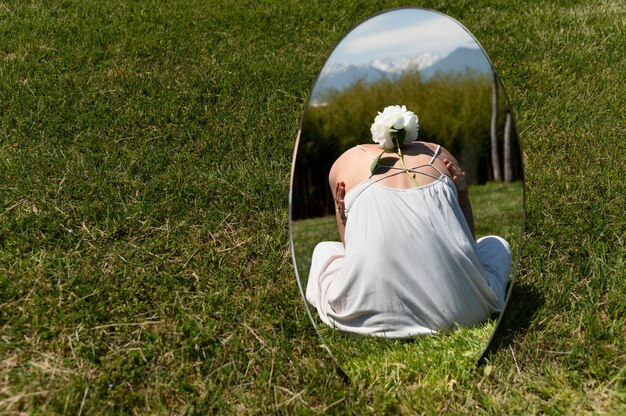 Young woman posing with peony flower in the mirror on the grass outdoors