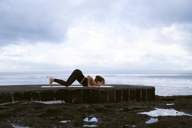 Молодая женщина практикует йогу на красивом пляже на рассвете. Голубое небо, океан, волны, близость к природе, единение с природой.