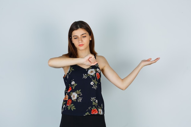 Young woman pretending to look at watch on her wrist, spreading palm aside in blouse, skirt and looking pensive. front view.