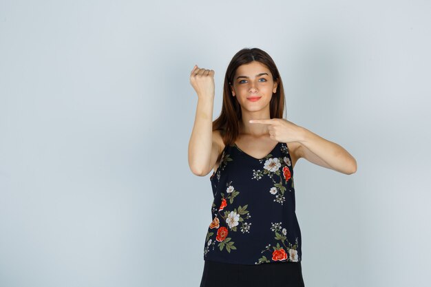 Young woman pretending to point at watch on her wrist in blouse, skirt and looking cheerful , front view.