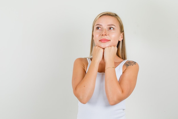 Young woman propping chin with fists in white singlet and looking hopeful.