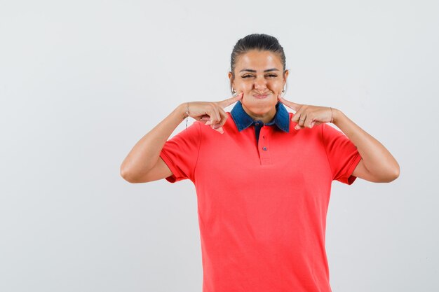 Young woman puffing cheeks, holding index fingers near mouth in red t-shirt and looking pretty. front view.