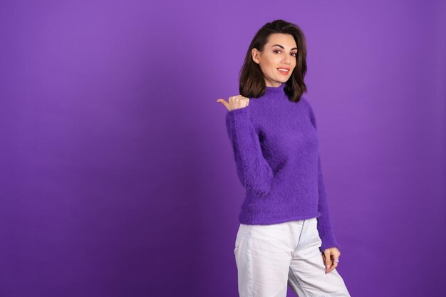 Молодая женщина в фиолетовом мягком уютном свитере на фоне милой, весело улыбаясь, в приподнятом настроении, указывает пальцем влево на пустое место