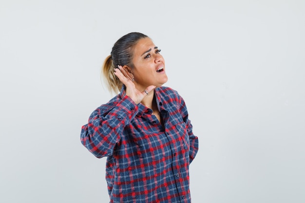 Young woman putting hand near ear and trying to listen in checked shirt and looking focused. front view.