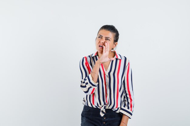 Young woman putting hand near mouth and calling someone in striped blouse and looking curious,