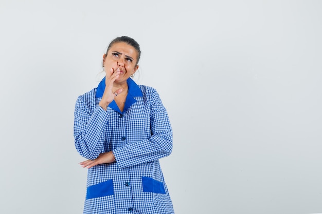 Young woman putting hand near mouth, standing in thinking gesture in blue gingham pajama shirt and looking pensive. front view.