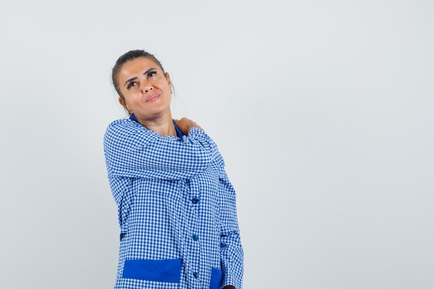 Young woman putting hand on shoulder, having shoulder pain in blue gingham pajama shirt and looking tired. front view.