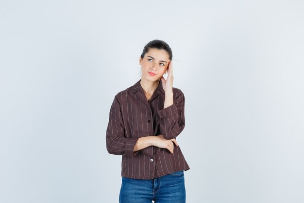 Young woman putting hand on temple, thinking about something in striped shirt, jeans and looking pensive. front view.