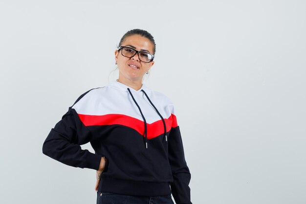 Young woman putting hand on waist in sweater, black jeans and glasses and looking pretty , front view.