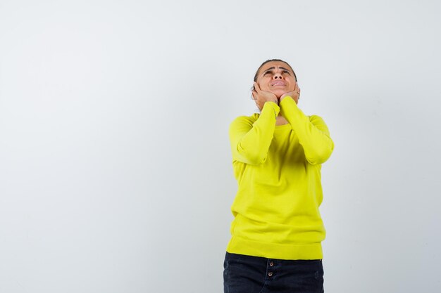 Young woman putting hands on cheeks, looking above in yellow sweater and black pants and looking happy 