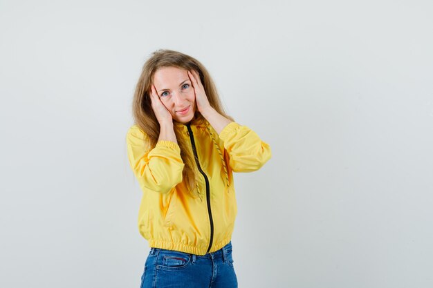 Молодая женщина кладет руки на лицо в желтой куртке бомбер и голубых джинсах и выглядит очаровательно, вид спереди.