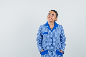 Бесплатное фото Молодая женщина кладет руки в карман, думая о чем-то в синей пижамной рубашке в клетку и задумчиво, вид спереди.