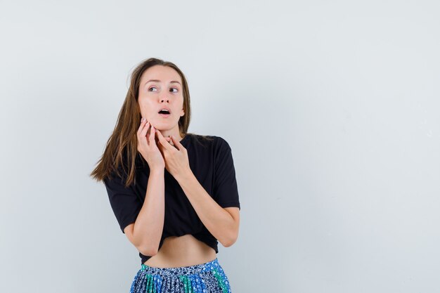 Giovane donna che mette le mani sul collo, distoglie lo sguardo in maglietta nera e gonna blu e sembra affascinante