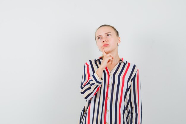 Free photo young woman putting her fingers on chin and looking upward in striped blouse and looking doubtful