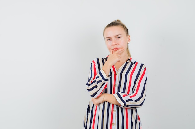 Young woman putting her fingers on chin and thinking about something in striped blouse and looking doubtful