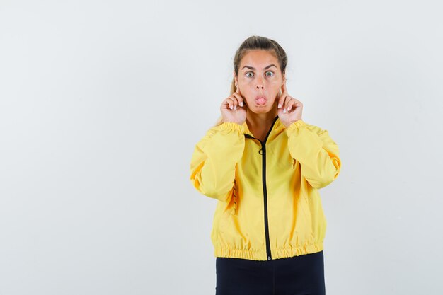 Young woman putting index fingers on ears while sticking tongue out in yellow bomber jacket and black pants and looking cute