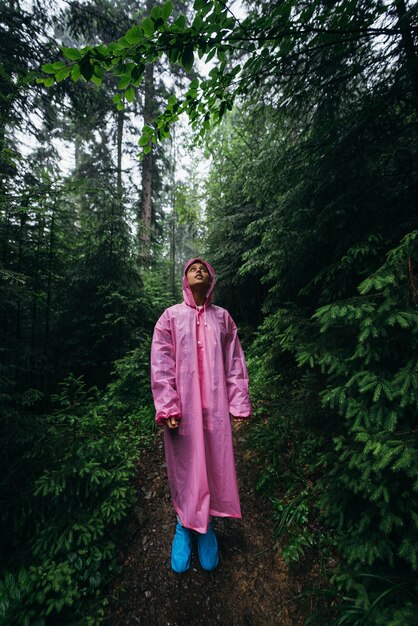 レインコートを着た若い女性が夕暮れの森の雨の中を歩く