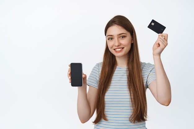 젊은 여성은 신용카드로 손을 들고 만족스럽게 웃으며 스마트폰 화면, 흰색으로 은행 업무를 위한 휴대전화 앱 인터페이스를 보여줍니다.