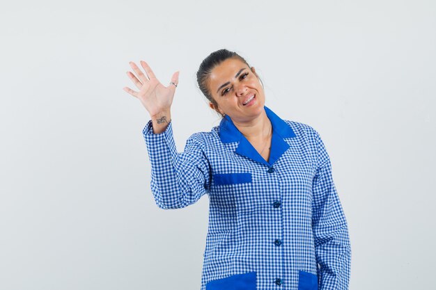 Young woman raising hand as greeting someone in blue gingham pajama shirt and looking pretty , front view.
