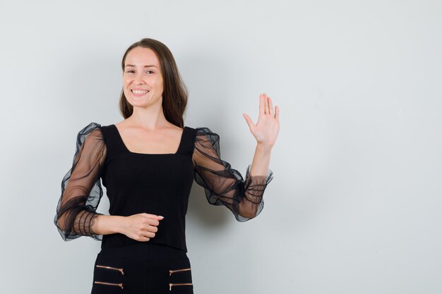 Young woman raising hand for greeting in black blouse and looking pleased 