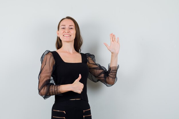 Young woman raising hand for greeting while showing thumb up in black blouse and looking cheerful. front view.