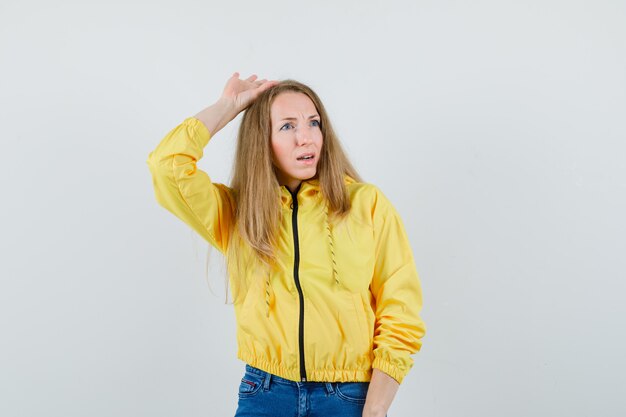 Young woman raising hand above head in yellow bomber jacket and blue jean and looking charming. front view.