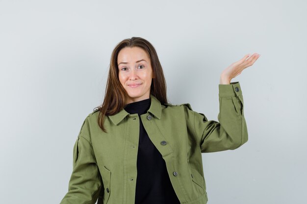 Young woman raising hand for showing something in green jacket and looking optimistic. front view.