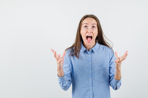 Young woman raising hands in aggressive manner in blue shirt and looking nervous.