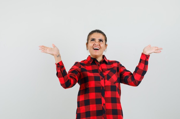 Young woman raising hands as holding something in checked shirt and looking glad. front view.