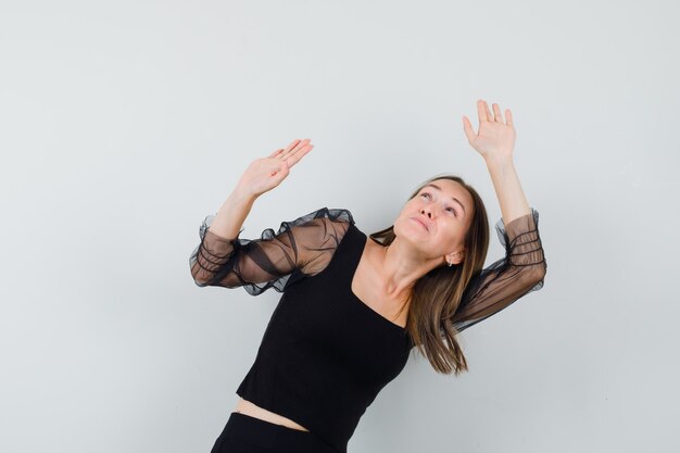 Young woman raising hands as holding something heavy in black blouse and black pants and looking tired. front view.