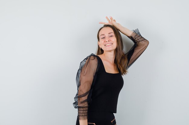 Young woman raising hands as showing bunny gesture in black blouse and black pants and looking amused , front view.