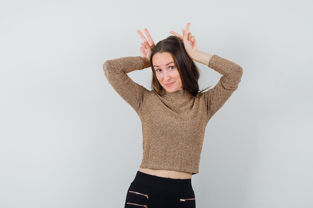 Young woman raising hands as showing bunny gesture in gold gilded sweater and black pants and looking happy. front view.