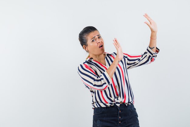 Young woman raising hands as to stop something in striped blouse and looking surprised.