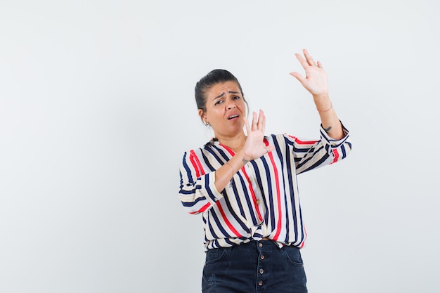 Young woman raising hands as stopping something in striped blouse and looking scared