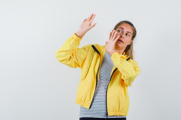 Young woman raising hands to defend herself in t-shirt, jacket and looking scared. front view.