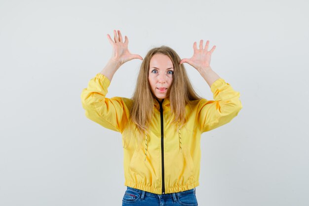 Young woman raising hands near head as showing deer horns in yellow bomber jacket and blue jean