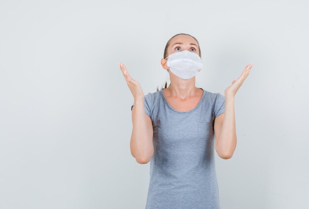 Young woman raising hands in praying gesture in grey t-shirt, mask front view.