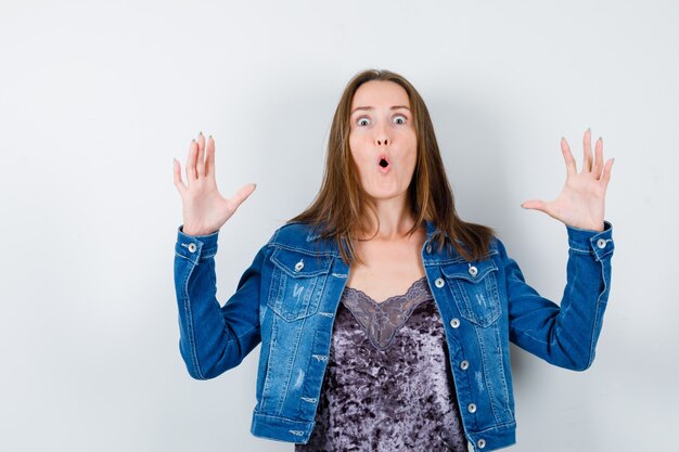 Young woman raising hands in surrender gesture in denim jacket, dress and looking terrified , front view.