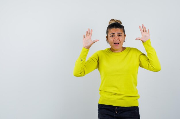 Young woman raising hands in surrender position in yellow sweater and black pants and looking serious