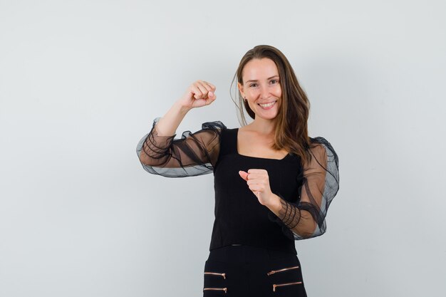 Young woman raising her fists in black blouse and looking confident. front view.