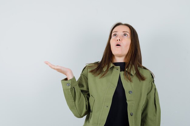 Young woman raising her hand with open palm in green jacket and looking surprised. front view.