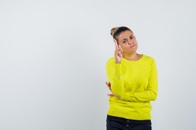 Young woman raising index finger in eureka gesture while holding hand on elbow in yellow sweater and black pants and looking sensible