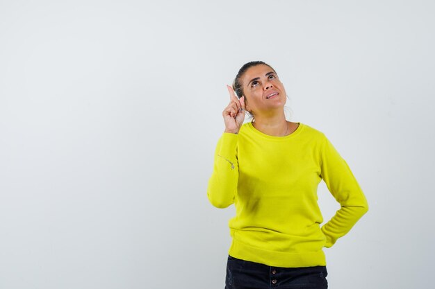 Young woman raising index finger in eureka gesture while holding hand on waist in yellow sweater and black pants and looking sensible