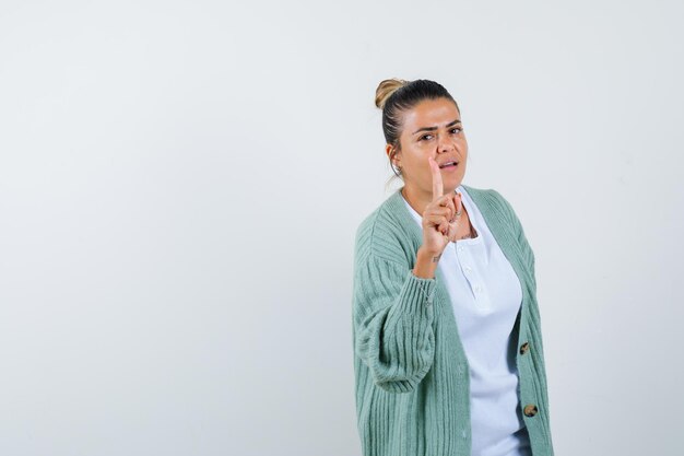 Young woman raising index finger in eureka gesture in white shirt and mint green cardigan and looking sensible