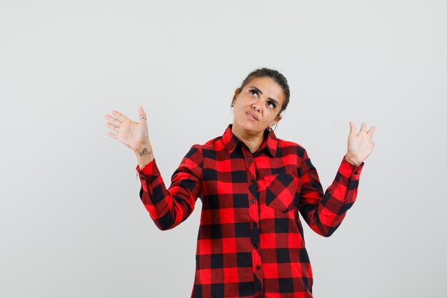 Young woman raising open palms while looking up in checked shirt front view.