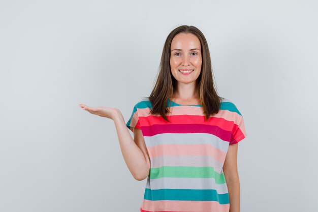 Young woman raising palm as catching something in t-shirt and looking glad , front view.