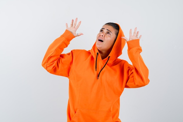 Free photo young woman raising palms in surrender gesture in orange hoodie and looking surprised